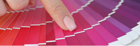Sample color chips in a range from red-oranges to purples printed into a swatch book, with pale skin hand pointing to a color in a shade of magenta