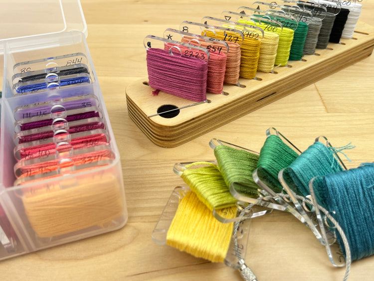 Clear acrylic bobbins are displayed in a bobbin box, on a cable ring, and in a wooden rack.
