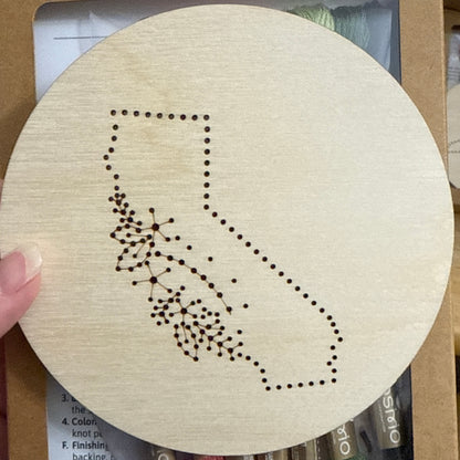 Wooden embroidery disk with a pre-patterned design of California. Packaging, embroidery floss, and instructions in the background.