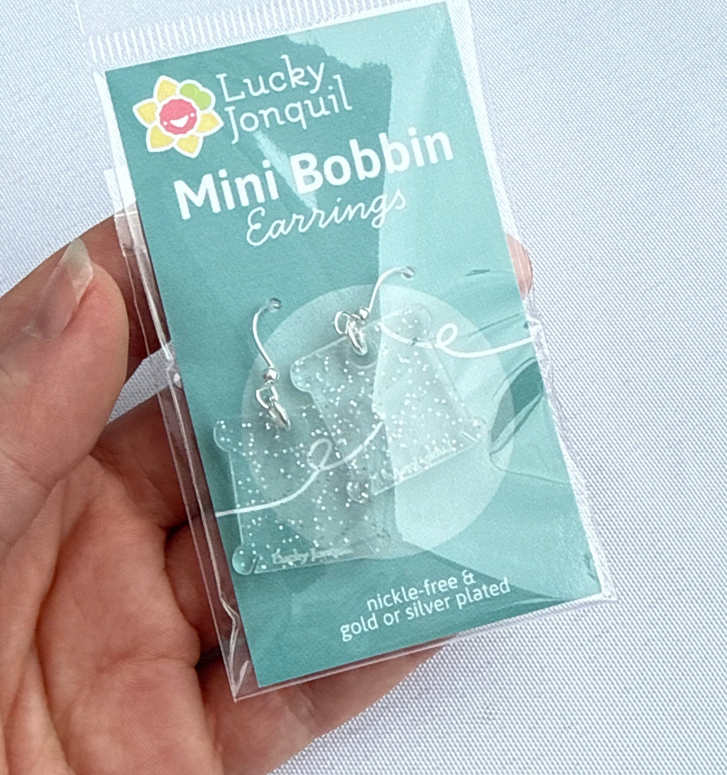 Packaged earrings in the shape of floss bobbins with a colorful holographic glitter and silver hooks