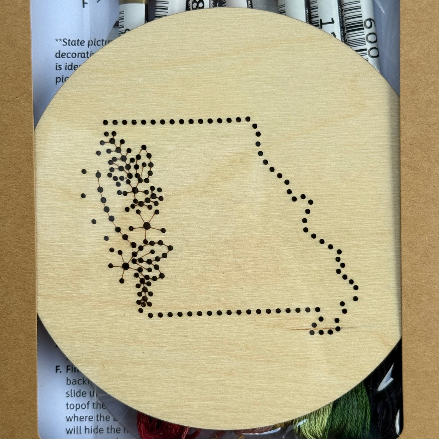 Wooden embroidery disk with a pre-patterned design of Missouri. Packaging, embroidery floss, and instructions in the background.