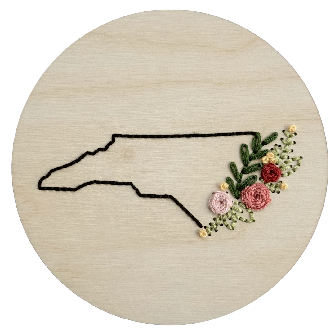 Wooden embroidery disk stitched with a black outline of North Carolina and a florals along the coast.