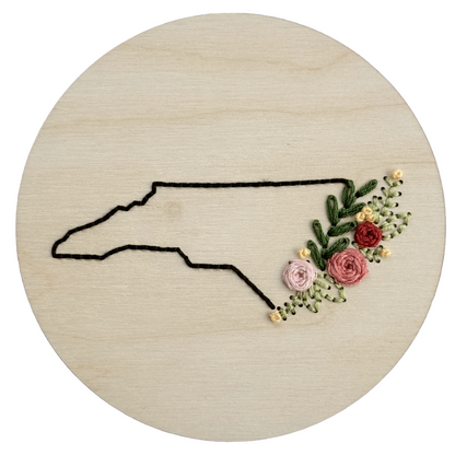 Wooden embroidery disk stitched with a black outline of North Carolina and a florals along the coast.