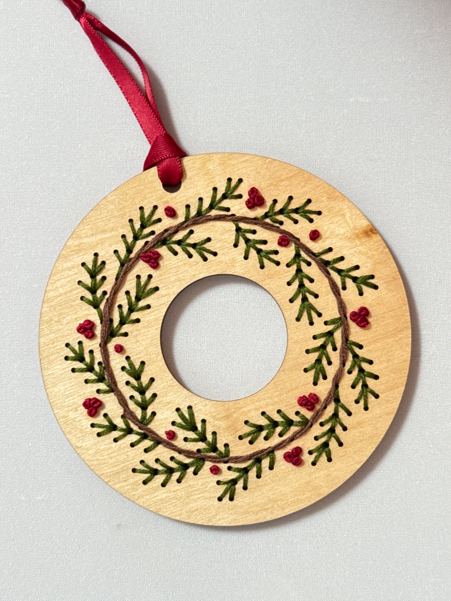Wooden wreath ornament with embroidered green leaves and red berries on a light gray background
